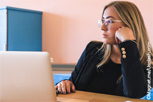 Picture: blonde woman with glasses sitting at a desk. Looking bored at a laptop screen.