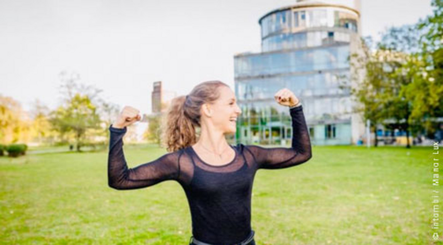 Young woman with black top in double biceps pose on a meadow in front of a futuristic building