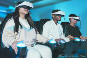 Three young people in casual clothes wearing virtual reality goggles sit in a row on cinema seats with blue illuminated buzzers in the armrests.