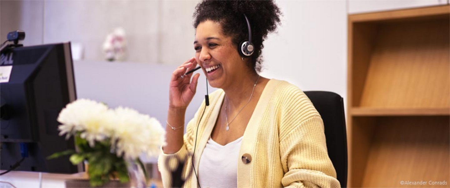 Lächelnde Mitarbeiterin des WiSSPo mit einem Headset in gelber Strickjacke an ihrem Arbeitsplatz.