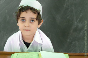 Brown haired young boy, sitting at a schooldesk, wearing a takka looking towards the camera.