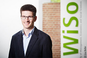 Portrait of WiSo Prof. Dr Clemens Kroneberg in a suit and wearing glasses. He stands in front of a brick wall and the WiSo Slogan on the right side.