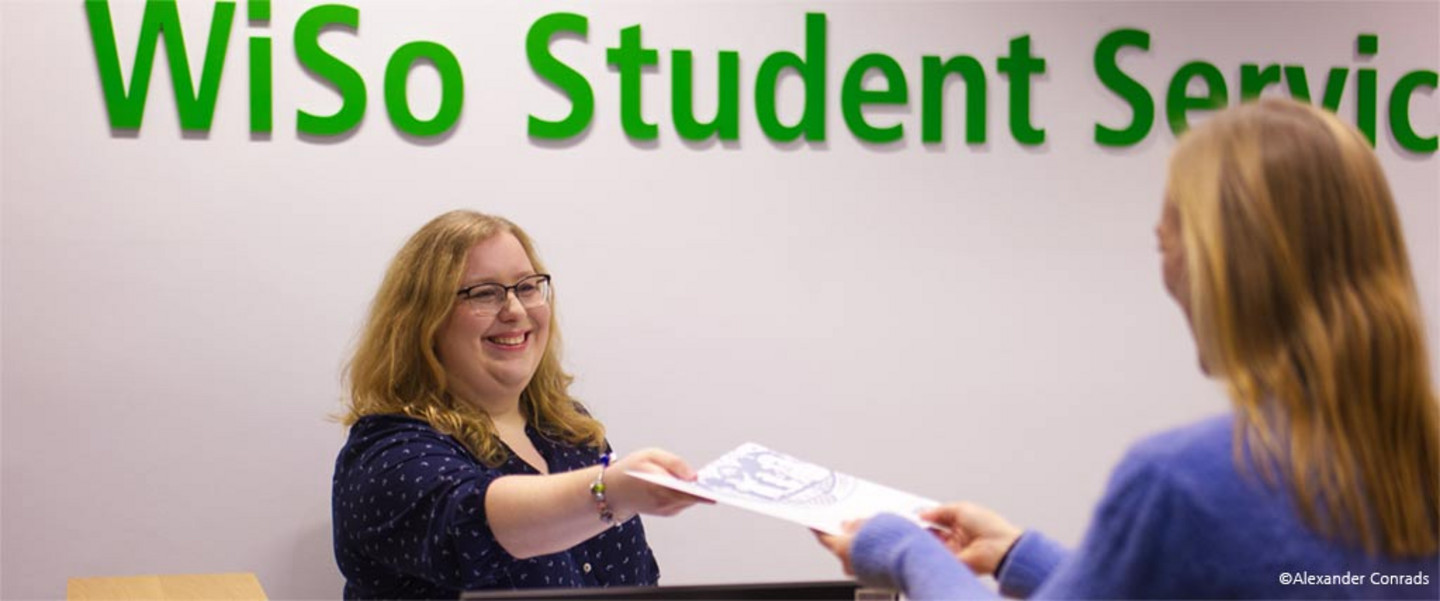 Eine WiSSPo-Mitarbeiterin reicht freundlich lächelnd einer Studierenden eine Dokumententenmappe mit dem Siegel der Universität zu Köln über den Tresen.