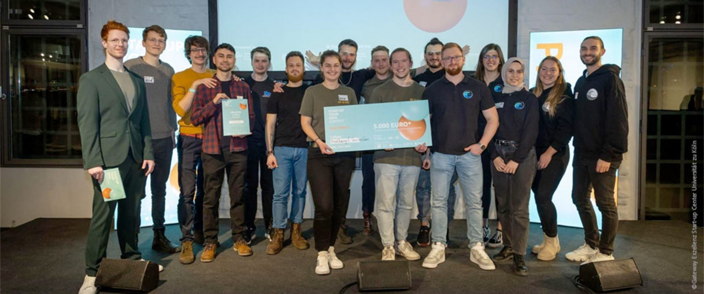 Winners of the fourth round of the Startup Your Idea Contest. Moderator Andreas Klein on stage with the 15 members of the two winning teams holding cheques and certificates.