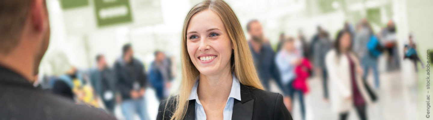Eine lächelnde Beraterin der Wirtschafts- und Sozialwissenschaftlichen Fakultät der Universität zu Köln in Businesskleidung steht in einer belebten Messehalle und spricht mit einem unscharf dargestellten Mann im Vordergrund. Im Hintergrund sind viele Menschen zu sehen, die durch die Halle gehen.