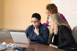Three women, two sitting at a desk with a laptop on it. Another standing behind them. Looking concentrated at the laptop screen.