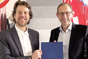 Jun.-Prof Raoul van Maarseveen und WiSo-Dekan Prof. Ulrich Thonemann Ph.D., lächelnd vor einem abstrakten Gemälde, halten gemeinsam eine blaue Mappe mit Logo der Universität zu Köln in die Kamera