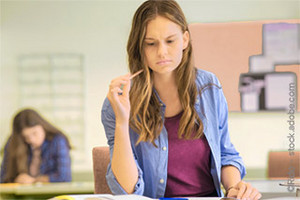 Young woman sitting at a desk in a seminar room, holding a pencil and looking thoughtfully at an open book, with another student blurred in the background.