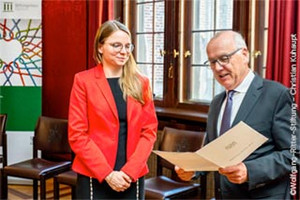 Digitalisation expert Dr Katharina Drechsler in a red jacket receives an award certificate from an older man in a suit in a formal setting.