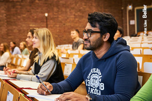 Studierende in einem leicht ansteigenden, mäßig besetzten Hörsaal mit einer Ziegelwand im Hintergrund schauen in guter Stimmung nach links aus dem Bild.