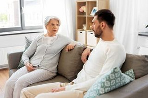 Eine ältere Frau mit weißen Haaren und ein jüngerer Mann sitzen auf der Couch. Sie sind einander zugewandt und schauen sich an, wie in einem Gespräch. Die Couch ist braun mit türkisfarbenen Kissen darauf. 