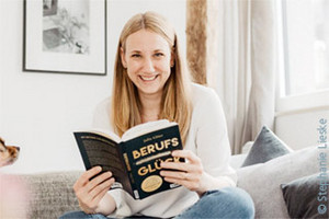 Jana Kielwein sitzt im Schneidersitz auf einem Sofa und schaut lächelnd auf, von einem Buch mit dem Titel "Berufsglück".