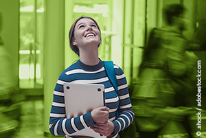 Eine junge Frau mit einem Laptop in der Hand. Sie hat einen grün weiß gesteiften Pullover an und braune Haare. Sie schaut lächeln nach oben. Der Hintergrund ist in grün hinterlegt. Man sieht Personen und einen Teil eines Gebäudes von innen. 