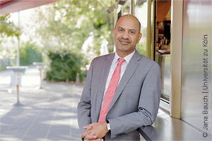 Prof. Dr. Joybrato Mukherjee in hellgrauem Anzug im Sonnelicht unter einer Markise an die Taschenablage eines Kaffeestands gelehnt gelehnt