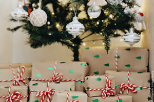 A festively decorated Christmas tree with white and silver ornaments and small figurines. The tree is lit with string lights. At the base, numerous gifts wrapped in brown paper with red and white twine are stacked. The background shows a bright wall and a cosy atmosphere.