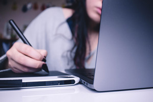 Eine Frau sitzt an einem Schreibtisch mit einem Laptop und schreibt mit einem Stift auf einem Tablet