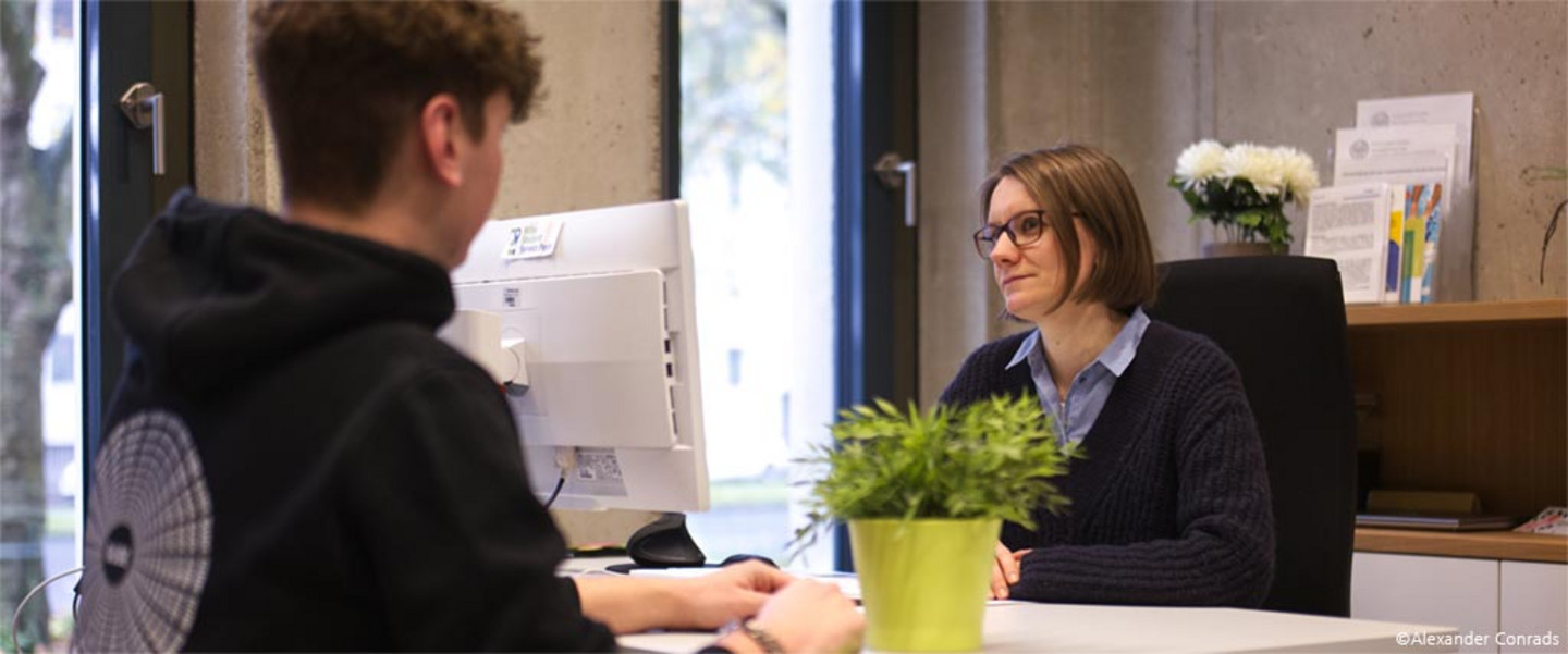 Eine Frau mit Brille an einem Schreibtisch in einem modernen Büro hört dem Anliegen eines Studierenden zu.