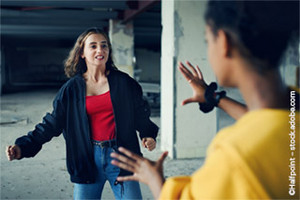 Zwei Teenager-Mädchen in einer konfrontativen Haltung, eine wirkt aggressiv und die andere defensiv, in einem Innenbereich, möglicherweise in einem verlassenen Gebäude.