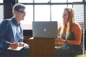 Blonde guy and woman, sitting next to each other talking. Between them stands a laptop.