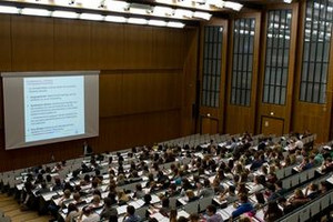 Bild: Ein voller Hörsaal an der Universität zu Köln. Vorne wird eine Präsentation gezeigt, die Student:innen hören zu. 