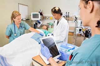Drei Krankenhausmitarbeiter*innen um ein Patientenbett (im Aufwachraum), davon eine Schwester mit einem tablet.