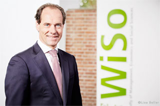 Professor Werner Reinartz, Ph.D. im Anzug vor einer Zeigelwand mit WiSo-Banner (Nahaufnahme)