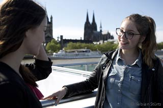 WiSo Buddy Programme - Two young women talking at an excursion boat railing in front of the Cologne Cathedral