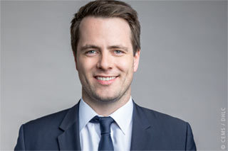 CEMS Alumni Alexander Schütt, Frontalansicht, lächelnd im Anzug, vor einem grauen Hintergrund.