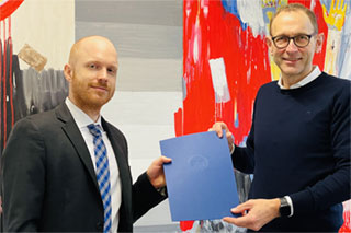 Professores Tobias Bauckloh und Ulrich Thonemann vor einem abstrakten Gemälde halten gemeinsam eine blaue Mappe 