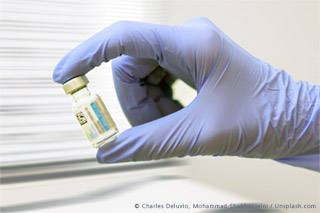 Eine HAnd in einem medizinischen Handschuh hält ein Impfstoffampulle vor einer medizinischen Liege