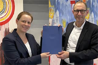 Handshake of Professores Paula Protsch and Ulrich Thonemann, jointly holding the certificate of appointment, in front of an abstract painting