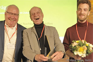 Reiner Bieck hands over the German Demography Award 2023 to Burghard Flieger (with the award) and Joschka Moldenhauer (with flowers) in front of a yellow presentation slide. 