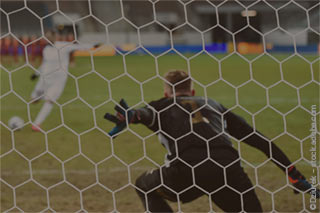 A football player taking a penalty kick with a goalkeeper preparing to save, seen through the net from behind the goal.
