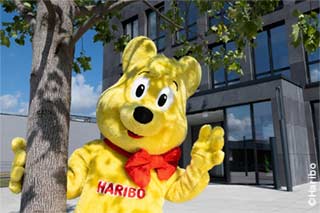 The HARIBO Goldbear standing under a tree in front of the HARIBO headquarters in Grafschaft, illustrating a cooperation with the University of Cologne's WiSo Faculty.