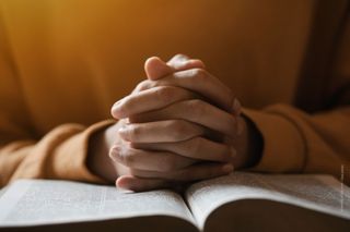 A person with folded hands, the hands are placed on an open book. The person ist wearing a yellow pullover. 