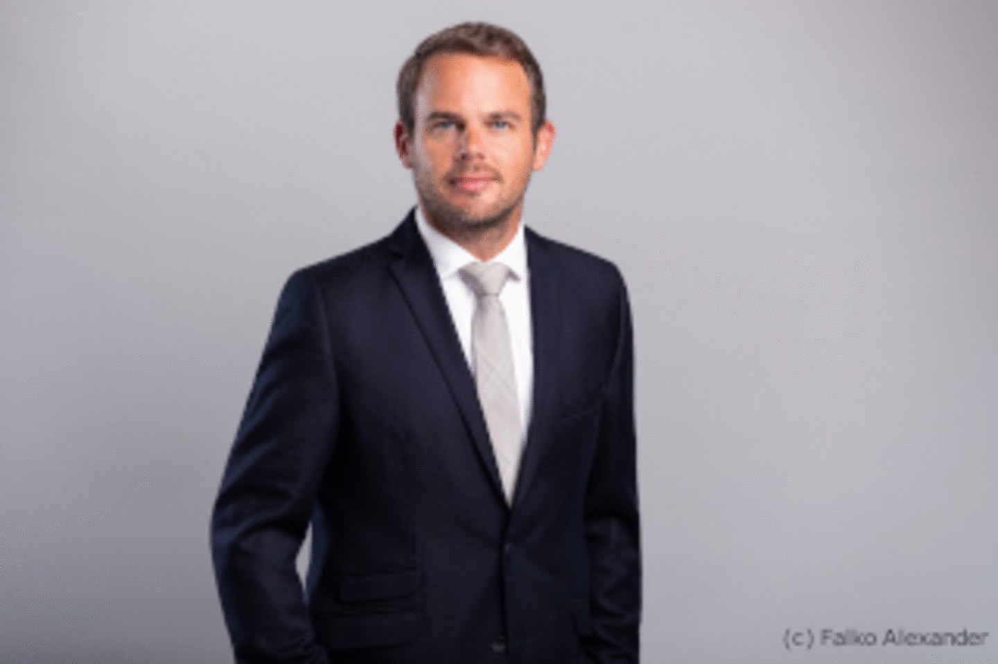 Portrait of Florian Sonnenburg in a black suit in front of a grey wall