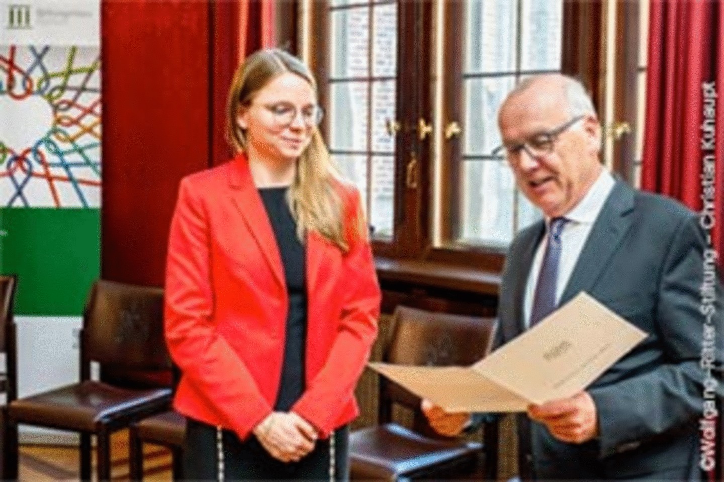 Digitalisierungsexpertin Dr. Katharina Drechsler in einer roten Jacke erhält ein Auszeichnungszertifikat von einem älteren Mann in einem Anzug in einem formellen Rahmen.