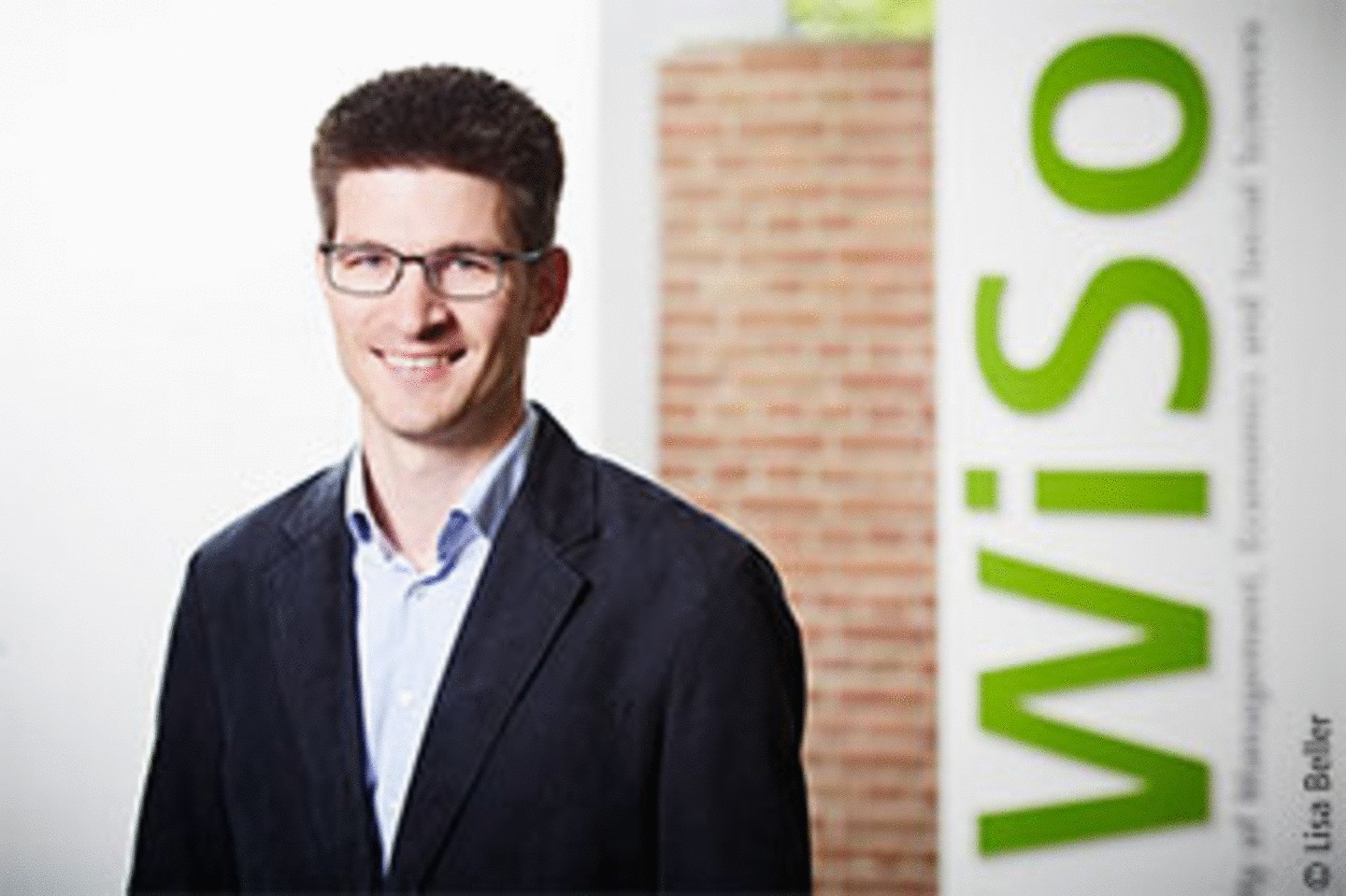 Portrait von WiSo-Professor Dr. Clemens Kroneberg im Anzug mit Brille vor einer Backsteinmauer und dem WiSo Schriftzug im Hintergrund. 