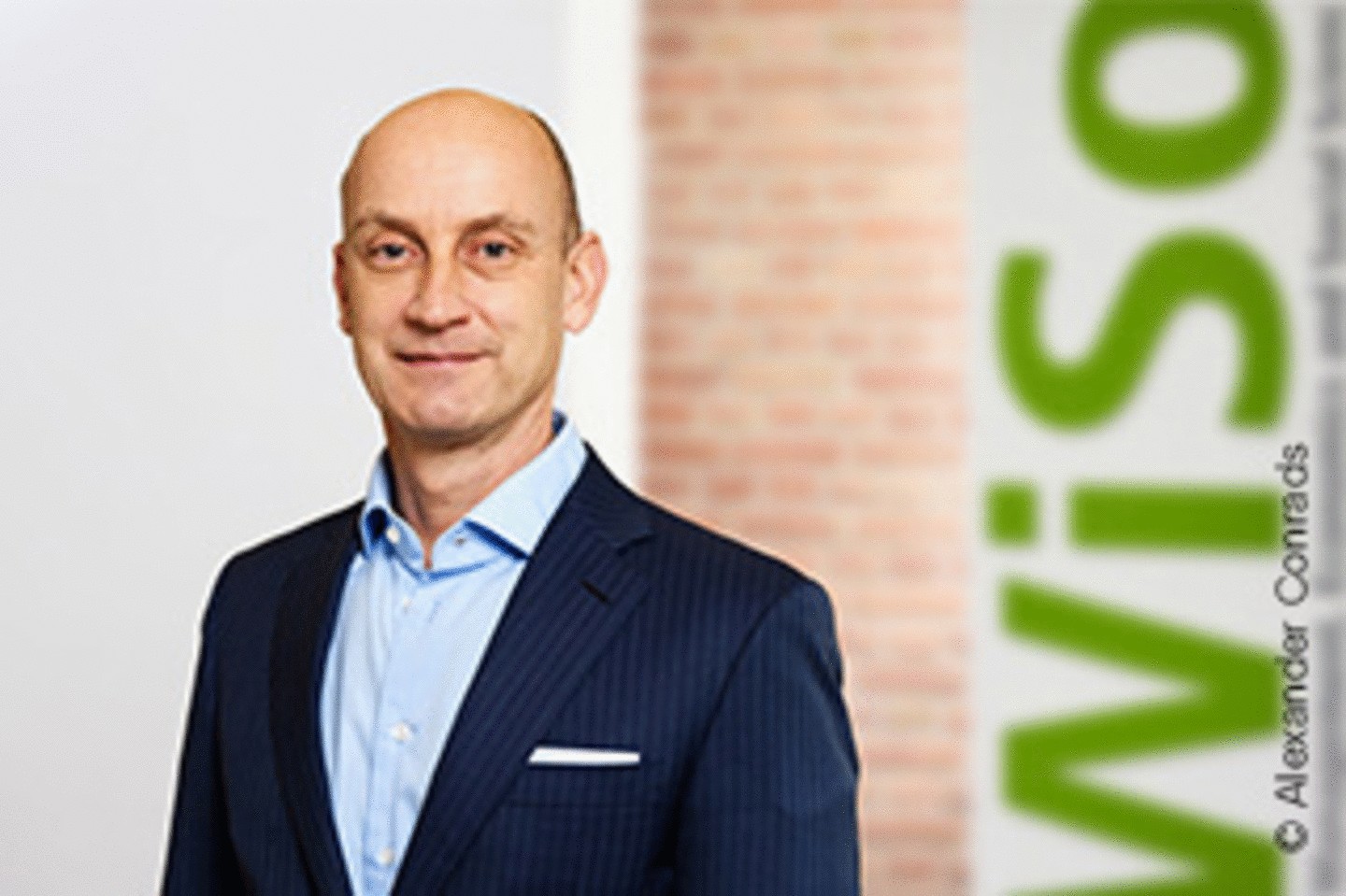 WiSo Professor Marc Fischer in a blue suit. Smiling towards the camera. A stone brick wall in the background and the WiSo Faculty logo and slogan in the background on the right.