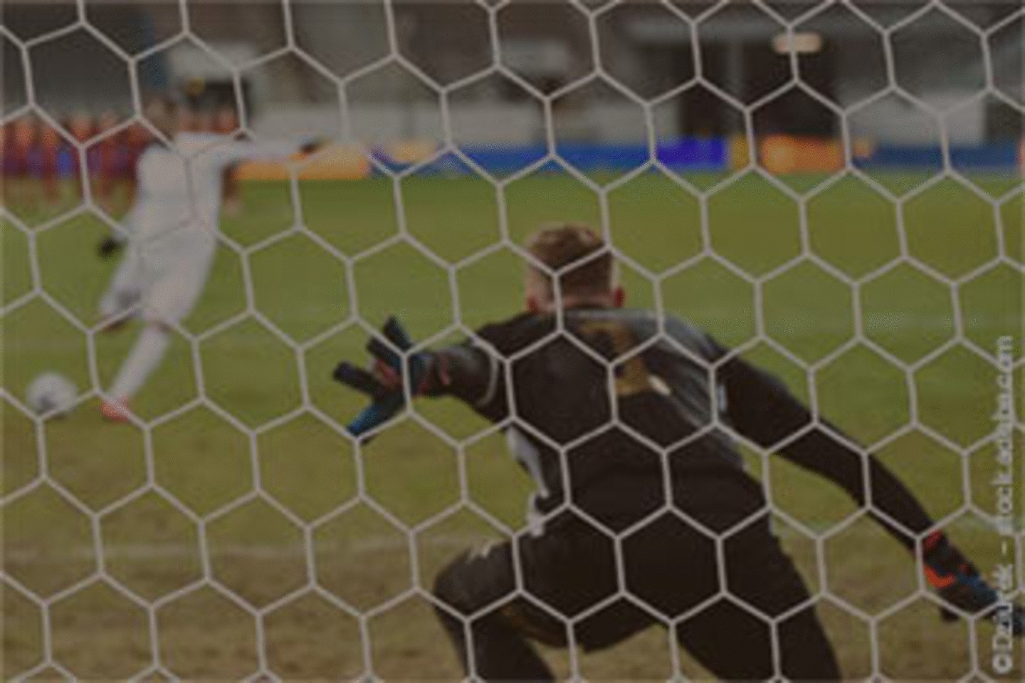 Ein Fußballspieler schießt einen Elfmeter, während der Torwart sich auf die Abwehr vorbereitet, durch das Netz von hinten gesehen.